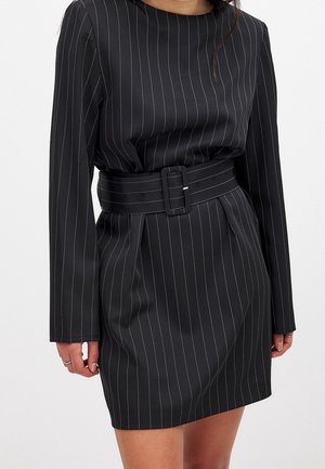 Robe noire à fines rayures blanches verticales, manches longues, et une large ceinture à la taille. Le tissu semble lisse et légèrement brillant.
