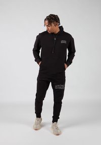 Felpa nera con zip e pantaloni della tuta abbinati con stampa "WRONG FRIENDS". Tessuto morbido, vestibilità rilassata, polsini a costine e sneakers con design testurizzato.