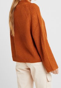 Kvinna med långt hår som bär en orange ribbstickad tröja och ljusbeige byxor med sidoficka, stående mot en enfärgad bakgrund.