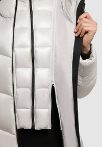 Lys grå pufferjakke med en glat overflade, sorte lynlåse og et paneldesign med varierende puffstørrelser, der viser en lynlås fortil.