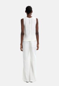 Biała lniana bluzka bez rękawów z postrzępionymi brzegami, zapinana na guziki z tyłu, w połączeniu z szerokimi lnianymi spodniami w kolorze białym, posiadającymi boczną kieszeń.