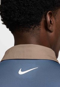 Nike polo krekls ar zilu korpusu, brūnu apkakli un baltu logotipu. Izceļas ar teksturētu audumu un pieguļošu dizainu. Tuva ainava no aizmugures.