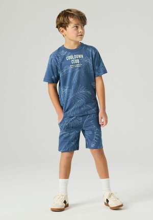 Junge steht und trägt ein blaues passendes T-Shirt mit tropischem Blattmuster und dem Text "COOL DOWN CLUB" sowie Shorts, weiße Socken und beige Turnschuhe.