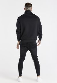 Maglione nero a mezza zip con collo alto, maniche raglan e polsini a coste, abbinato a jeans neri slim-fit e sneakers bianche.