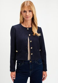 Blondynka w granatowej, fakturowanej kurtce z złotymi guzikami, beżowej bluzce i ciemnoniebieskich dżinsach, stojąca na tle białym.