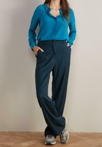 Turquoise button-up blouse with a v-neck and long sleeves, paired with dark blue wide-leg trousers and gray sneakers. Smooth fabric texture.