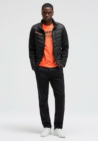 Chaqueta negra acolchada con cuello alto, camiseta gráfica naranja, pantalones oscuros y zapatillas blancas. La chaqueta tiene un cierre de cremallera y detalles de logotipo.