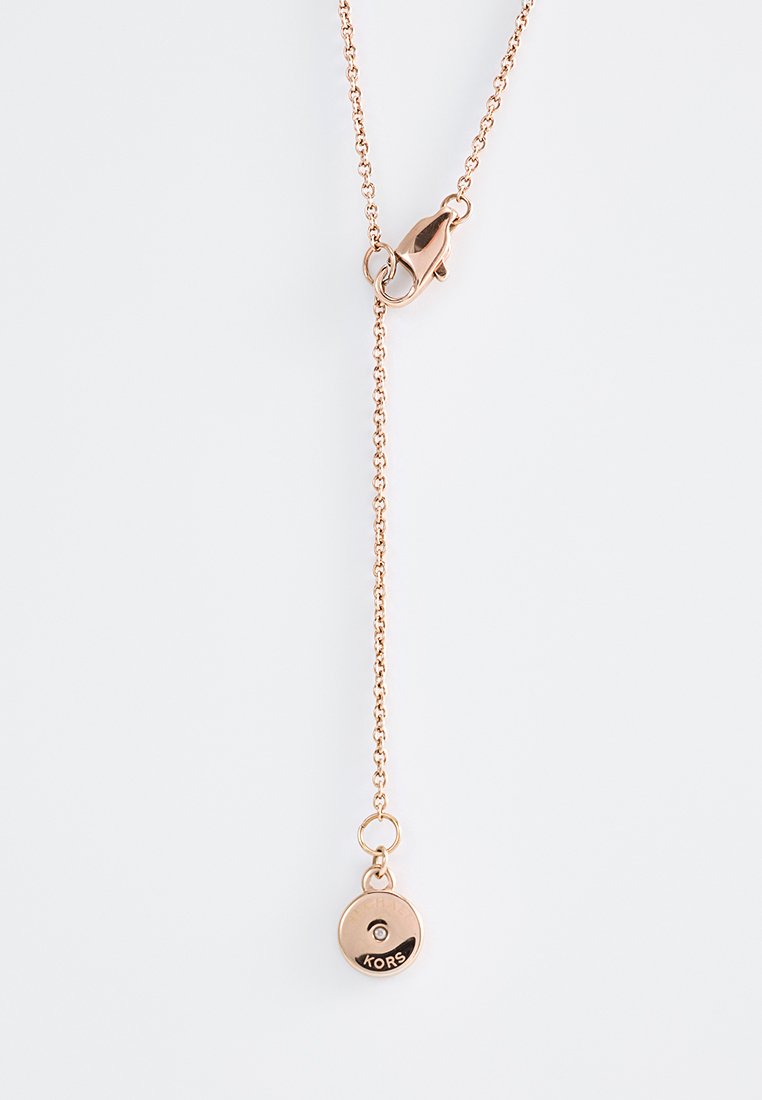 Collier en chaîne en or rose avec fermoir à homard et petit pendentif rond gravé avec "KORS" sur fond blanc.