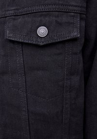 Chemise en denim noir avec une poche à rabat arrondi, des coutures foncées et un bouton en métal avec le logo de la marque. Surface du tissu texturée.