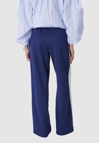 Pantalon large bleu avec une texture lisse, présentant une rayure latérale blanche contrastante. Le matériau semble léger et extensible.