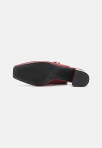 Chaussure à talon aigu rouge avec une tige lisse, un bout carré, une semelle en caoutchouc noir et un petit accent sur le côté.