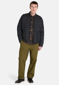 Chaqueta negra acolchada con cierre de botones a presión, llevada sobre una camisa de cuadros marrón, combinada con pantalones verdes y zapatos negros.