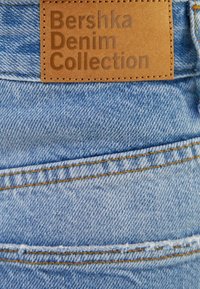 Ljusblå denimtyg med synliga sömnadsdetaljer. En rektangulär brunt läderetikett nära toppen med präglad text "Bershka Denim Collection."