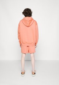 Korallfärgad hoodie med huva och ribbade ärmslut, tillsammans med matchande shorts. Tyget verkar mjukt med en jämn textur. Sneakers är vita med orangea och bruna accenter.