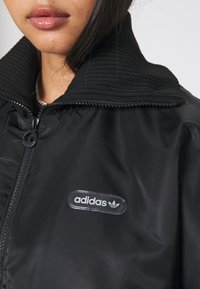 Veste matelassée noire avec un col côtelé, fermeture éclair à l'avant et un logo Adidas noir et blanc bien visible sur la poitrine. Texture lisse et brillante.
