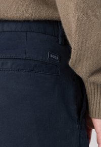 Gros plan d'une personne portant un pantalon bleu marine avec une petite étiquette "BOSS" près de la poche arrière et un pull en tricot beige.