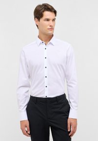 Camicia bianca a maniche lunghe con colletto strutturato, dotata di bottoni neri e una texture liscia, abbinata a pantaloni scuri.