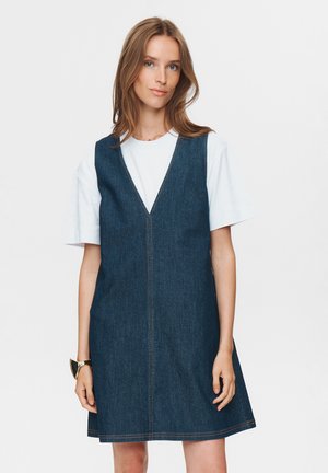 Abito in denim con scollatura a V, forma a campana e cuciture a vista. Stilizzato sopra una maglietta bianca, offre un look casual e a strati.