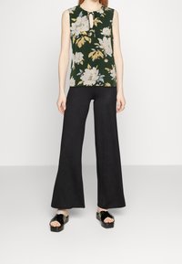 Kvinna i ärmlös mörkgrön blommig blus, svarta vida byxor och svarta plattformssandaler, stående mot en neutral bakgrund.