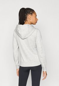 Γυναίκα με πλεξούδες, γυρισμένη πλευρικά, φοράει ανοιχτό γκρι φούτερ με κουκούλα και μαύρα κολάν, ενάντια σε απλό φόντο.