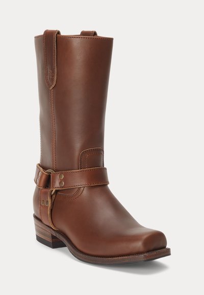 Kõrge pruun nahast läänepoolsed saapad rihma ja metallnööpide detailiga, virnastatud puidust konts ning ümar nurgaga ruudukujuline ninaosa.