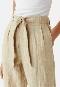 Beige lněné kalhoty s páskem na zavazování, předními sklady a postranními kapsami. Materiál má texturovaný povrch.