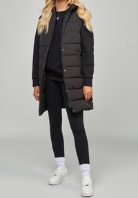 Black puffer vest with a hood, featuring a button closure and quilted design. Worn over a dark sweatshirt and black leggings with white sneakers.