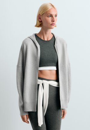 Femme blonde portant un haut court gris, des leggings assortis, un sweat à capuche gris clair zippé et un sweat blanc noué autour de la taille.