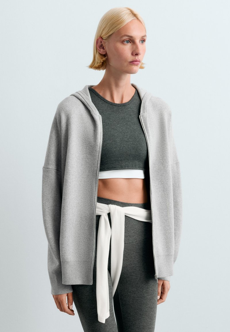 Femme blonde portant un haut court gris, des leggings assortis, un sweat à capuche gris clair zippé et un sweat blanc noué autour de la taille.