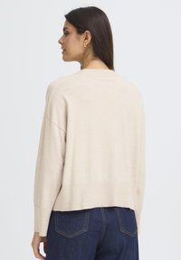 Bež pulover z okroglim izrezom in padajočimi rameni, pleten. Rebrast rob in manšete. Na hrbtni strani ima nedokončan rob, ki dodaja detajl.