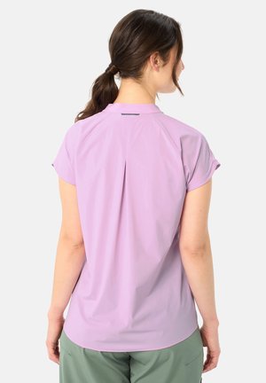 Donna con capelli scuri raccolti in una coda di cavallo, che indossa una maglietta lilac a maniche corte e pantaloni verdi, vista di spalle su uno sfondo bianco neutro.