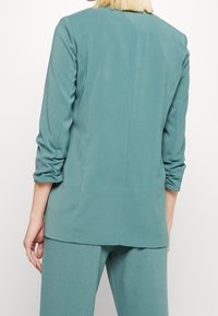 Op maat gemaakte, teal blazer met gerimpelde mouwen, gemaakt van zachte stof. Heeft een rechte snit en achternaaddetails, waardoor een soepele silhouet ontstaat.