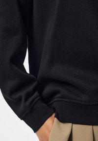 Personne portant une chemise noire à manches longues, la main partiellement dans la poche d'un pantalon beige, sur un fond uni.
