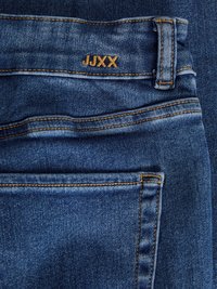 Jeans de denim en azul profundo con costuras naranjas, que cuentan con una etiqueta de marca "JJXX" en la cinturilla y un bolsillo trasero con detalles de costura.
