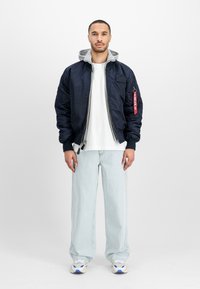 Hombre de pie vestido con una chaqueta bomber azul marino sobre una sudadera gris, camiseta blanca, jeans de pierna ancha en azul claro y zapatillas blancas, sobre un fondo blanco.
