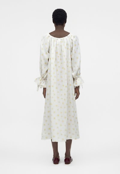 Γυναίκα που στέκεται πλάτη, φορώντας ένα λευκό μίντι φόρεμα με κίτρινο λουλουδάτο σχέδιο και μακριά φουσκωτά μανίκια, συνδυασμένο με σκούρα κόκκινα επίπεδα παπούτσια.