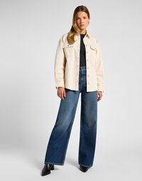 Woman wearing a cream button-up shirt over a black top, wide-leg blue jeans, and black ankle boots, standing against a plain light background.
