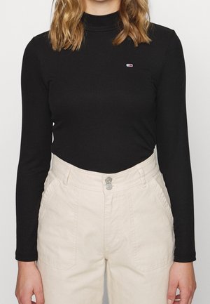 Frau trägt ein eng anliegendes schwarzes Langarmshirt mit kleinem Logo auf der Brust und hoch taillierte, cremefarbene Hosen mit Fronttaschen.