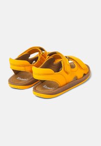 Keltaiset sandaalit, joissa on säädettävät velcro-hihnat, pehmustettu pohja ja teksturoitu ruskea ulkopohja. Sivujen aukot hengittävyyden lisäämiseksi.