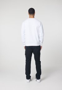 Uomo in piedi, di spalle, con indosso un maglione bianco, pantaloni cargo neri e sneakers bianche, su uno sfondo chiaro e uniforme.