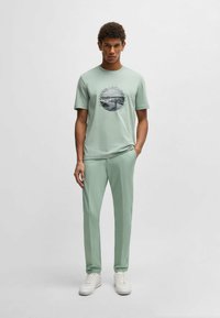 Jasnozielona koszulka z okrągłym nadrukiem graficznym, w połączeniu z dopasowanymi spodniami i białymi trampkami. Gładka tekstura na całej powierzchni.