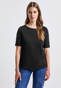 Femme aux longs cheveux bruns portant un haut noir à manches courtes et un jean bleu, se tenant devant un fond clair et uniforme.