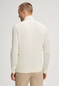 Witte kabelbrei trui met geribde kraag en manchetten, kenmerken textuurpatronen en een aansluitend ontwerp. De stof lijkt zacht en warm.