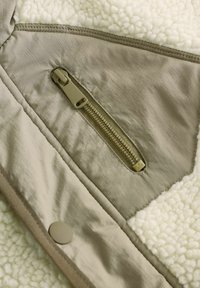 Leichte Jacke mit einer cremefarbenen Fleece-Fütterung, olivgrünem Außengewebe und einer Reißverschlusstasche mit beige abgesetzten Nähten.