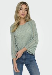 Top lavorato a maglia verde chiaro con una vestibilità ampia, caratterizzato da un motivo testurizzato, maniche larghe e scollatura tonda, abbinato a jeans in denim blu.