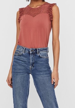 Bezrękawnik w kolorze rdzawej czerwieni z koronkowym karczkiem i falbankami przy rękawach, zestawiony z niebieskimi, wysokim stanem dżinsami na stojącej osobie.
