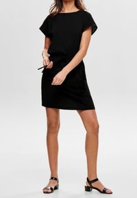 Schwarzes Kleid mit kurzen Ärmeln, tailliertem Bund, lässigem Schnitt und Länge über dem Knie. Getragen mit schwarzen Riemchensandalen.