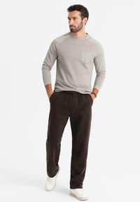 Camisa de manga larga gris claro con un bolsillo en el pecho, combinada con pantalones marrones oscuros y zapatillas blancas; ajuste casual con una textura suave.