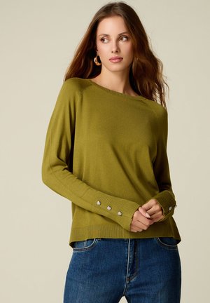 Donna con lunghi capelli castani che indossa un maglione verde oliva con dettagli a bottoni sulle maniche e jeans blu, che guarda di lato.
