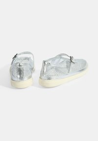 Silberne Netzschuhe mit runder Form, Gummisohlen und verstellbaren Riemen, die ein strukturiertes Muster über dem Obermaterial aufweisen.
